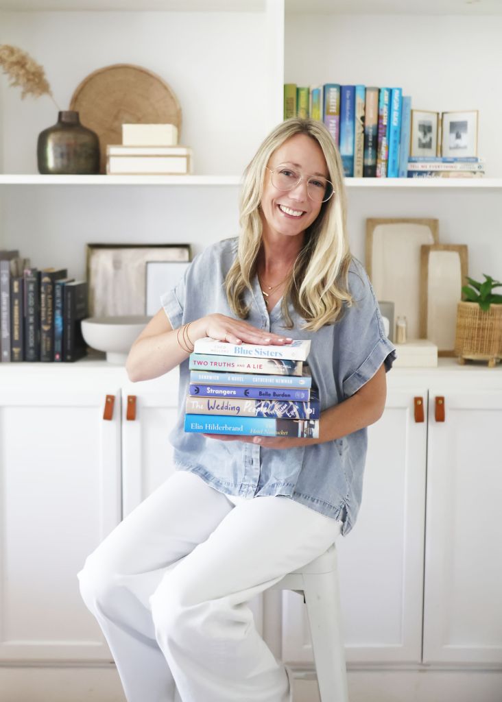 A woman with long blonde hair, wearing glasses and a blue shirt, smiles while sitting on a stool and holding a stack of books in her lap. In the background, there are shelves with books and decorative items.